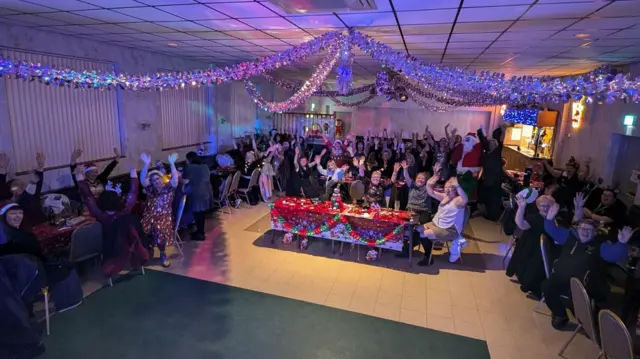 Dezenas de mulheres e uma pessoa vestida de Papai Noel acenam enquanto se sentam ao redor de mesas em uma sala de eventos decorada para o Natal.