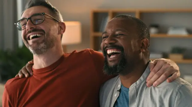 Dois homens abraçados e sorrindo em sala de estar