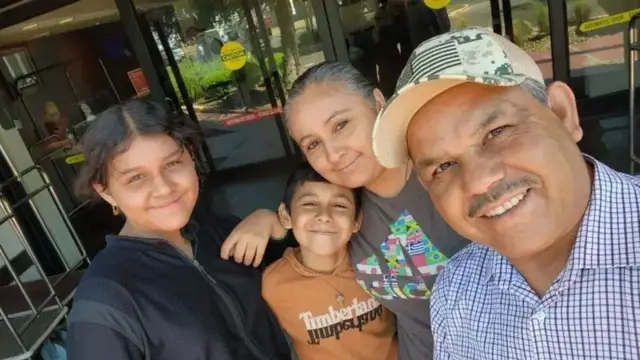 Da esquerda para a direita, uma menina vestindo um casaco preto de ziper está com o braço no ombro de um garoto vestindo uma camiseta marrom que está abraçado com uma mulher vestindo uma camiseta cinza. Na frente, um homem de camisa xadrez azul e branca e boné sorri