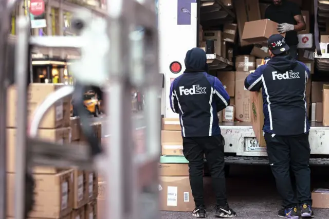 Dois trabalhadores da FedEx estão de costas, vestindo o uniforme da empresa que é escuro, e seguram caixas