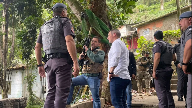 Três homens (um com farda de PM, outro com roupa social e outro com colete e fuzil) conversam em uma rua rodeada por casas e outros policiais