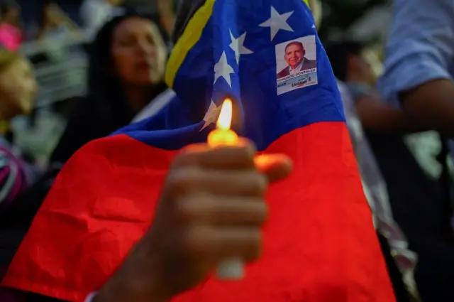 Uma pessoa com uma vela acesa, com o rosto coberto pela bandeira da Venezuela e segurando uma foto de Edmundo González