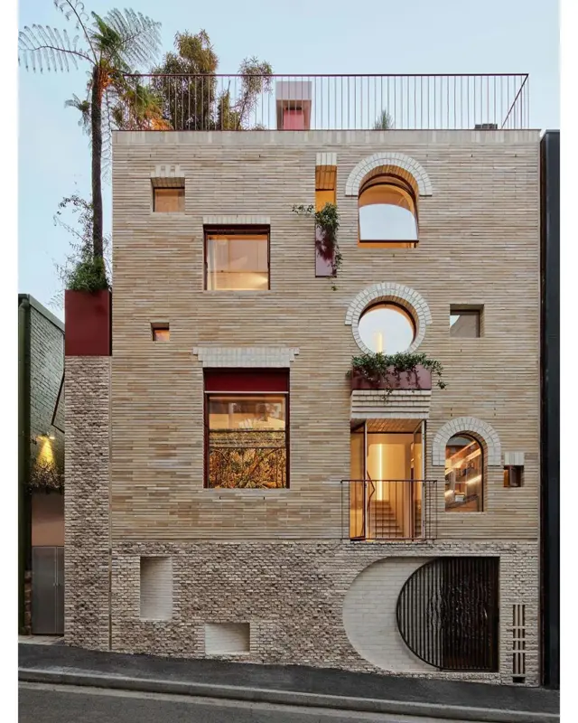 Fachada de casa; ela tem aparência excêntrica e janelas de tamanhos e espaçamentos irregulares, e um mosaico com materiais reciclados e tijolos quebrados.