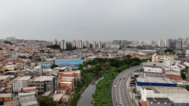 Um rio separa uma favela em São Paulo, no lado esquerdo, de galpões industriais e edifícios de São Caetano do Sul no lado direito; ao fundo, veem-se muitos prédios altos