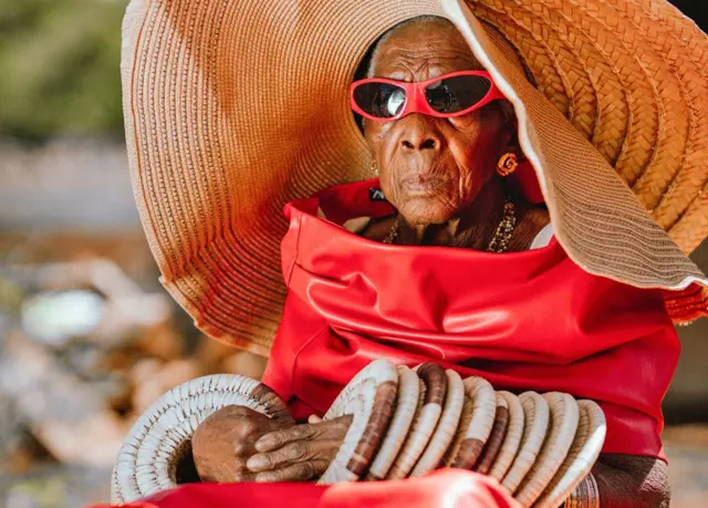 Margret Chola, sentada, parece séria enquanto usa um enorme chapéu de palha com pulseiras que se assemelham a tapetes de palha subindo por seus braços. Ela usa óculos de sol vermelhos e o que parece ser um vestido de couro vermelho.