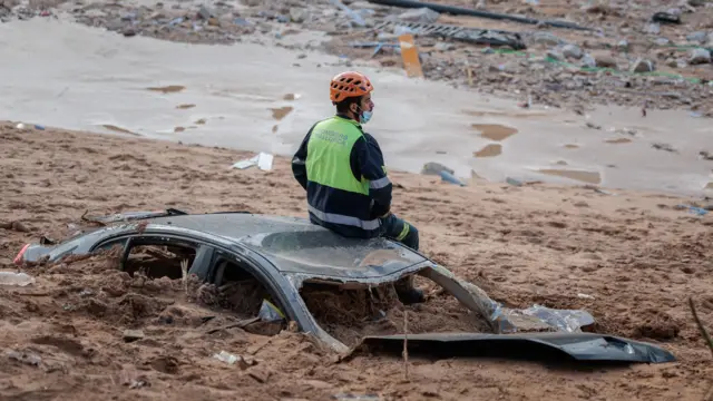 Foto mostra um socorrista sentado em cima de um carro enterrado na lama após inundações repentinas em Valência, Espanha