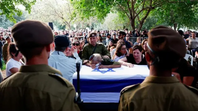 Funeral com várias pessoas. Caixão coberto com bandeira de Israel