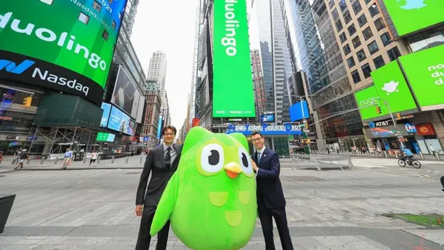 A foto foi feita na rua, com prédios ao redor de dois homens jovens vestidos com terno e gravata e uma coruja verde, símbolo do Duolingo, no meio deles. 