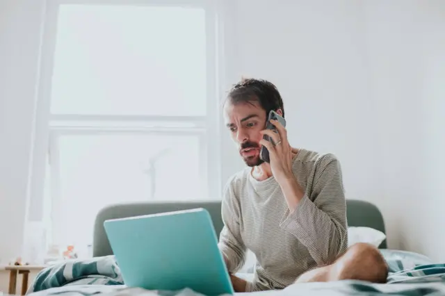 Homem branco segura o celular com cara de frustrado em frente a um laptop