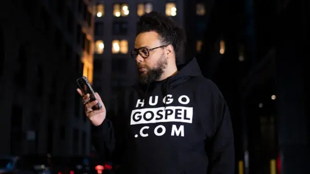 Gabriel Ribeiro em rua, durante a noite, olhando celular e usando moletom com estampa 'HugoGospel.com'