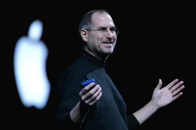 Steve Jobs, em uma blusa de gola alta preta durante uma apresentação da Apple, com o logotipo da Apple atrás dele