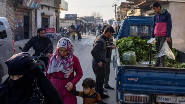 Pessoas caminhando por uma rua movimentada em um acampamento de refugiados em Idlib, com várias pessoas comprando verduras na traseira de um caminhão em 17 de dezembro