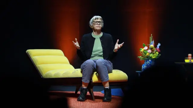 A foto mostra Philippa Perry, uma mulher de meia-idade, pele clara, cabelos curtos e franja, sentada em um divã amarelo gesticulando. 