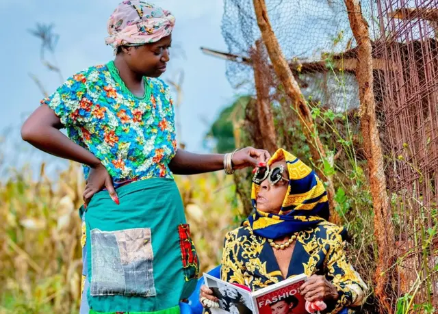 Diana Kaumba está de pé do lado de fora, vestindo roupas tradicionais zambianas, olhando para sua avó sentada, que usa uma jaqueta de grife estampada em azul-marinho e dourado, lenço e óculos de sol. Ela segura um livro com o título Hollywood Fashion, com Audrey Hepburn na capa.