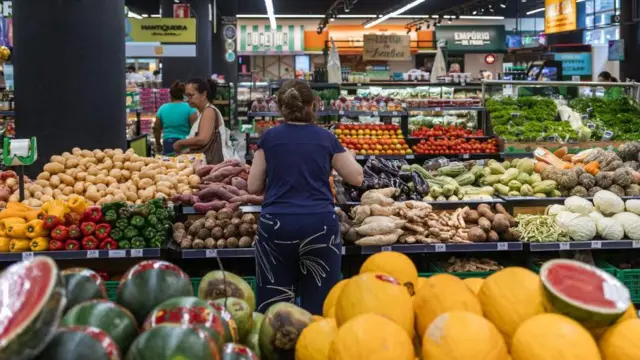 Uma mulher de costas escolhendo legumes na seção de vegetais de um supermercado com outras pessoas passando ao fundo