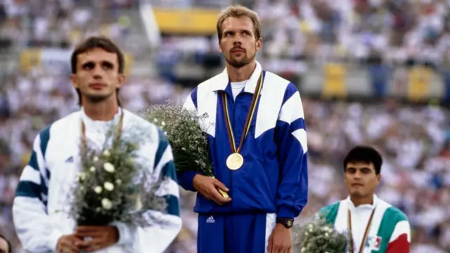 Andrey Perlov no topo pódio do Estádio Olímpico de Barcelona em 1992, com uma medalha de ouro no pescoço, segurando um buquê de flores — à sua esquerda e direita estão os competidores que ficaram em segundo e terceiro lugar na disputa