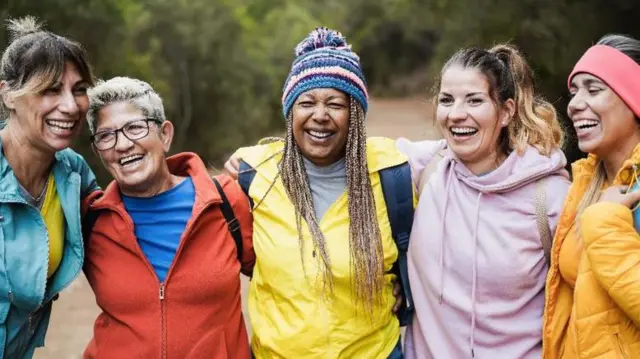 Amigas com idades variadas abraçadas e sorrindo em trilha de caminhada