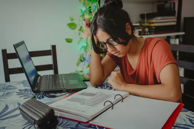 Mulher com óculos em uma mesa lendo anotações de um fichário, com um computador ao lado.