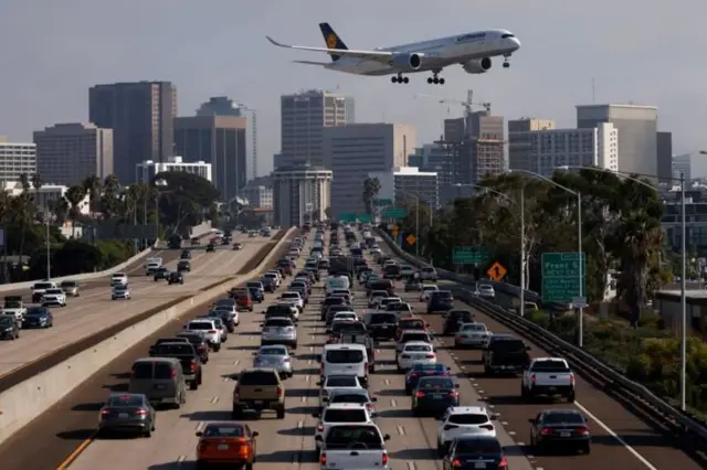 Avião passando perto da cidade, próximo a via engarrafada com prédios ao fundo