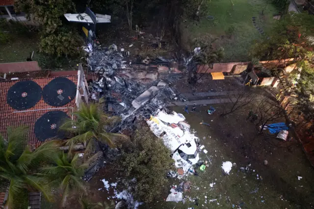 A imagem aérea mostra um gramado com destroços de um aviçao branco e do lado direito o telhado de uma casa