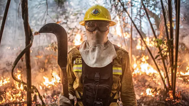 Brigadista usando máscara e foice para combater incêndios com floresta queimando ao fundo