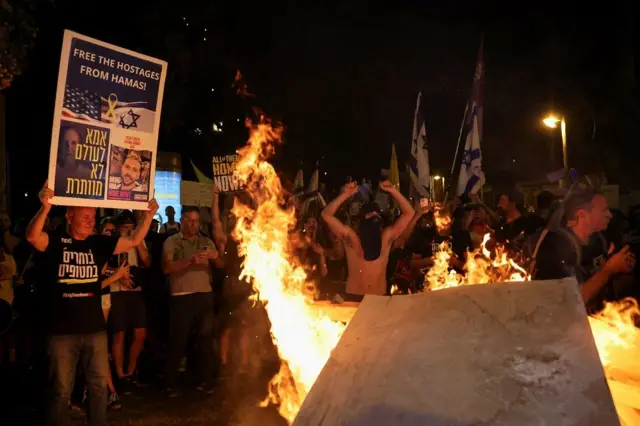 A foto mostra pessoas na rua, segurando cartazes e colocando fogo em algo, durante um protesto a noite. 