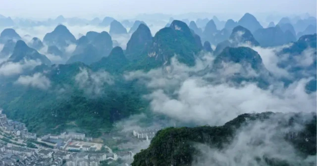 A imagem mostra uma paisagem natural composta por montanhas cobertas por vegetação, com cumes pontiagudos que emergem de uma névoa espessa, criando uma atmosfera etérea. Na parte inferior, há um pequeno aglomerado urbano com prédios e ruas, que contrasta com a imensidão das formações naturais. O céu tem tons azulados, reforçando o clima de serenidade e mistério da cena.