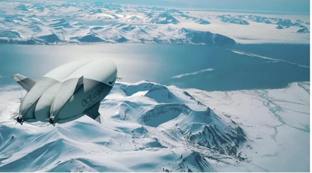 Dirigível branco sobrevoa paisagem de montanhas nevadas, com vasto lago ao fundo, sob céu claro e tons gelados