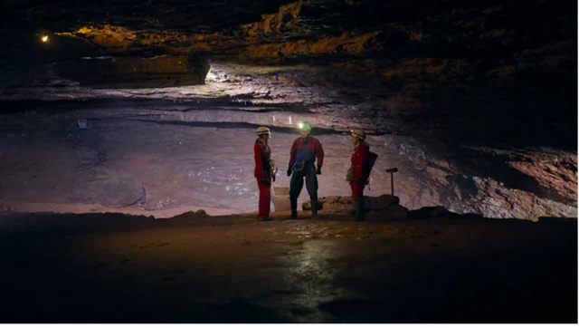 Três pessoas vestidas com roupas de exploração e capacetes com lanternas estão paradas em uma ampla caverna iluminada artificialmente. 