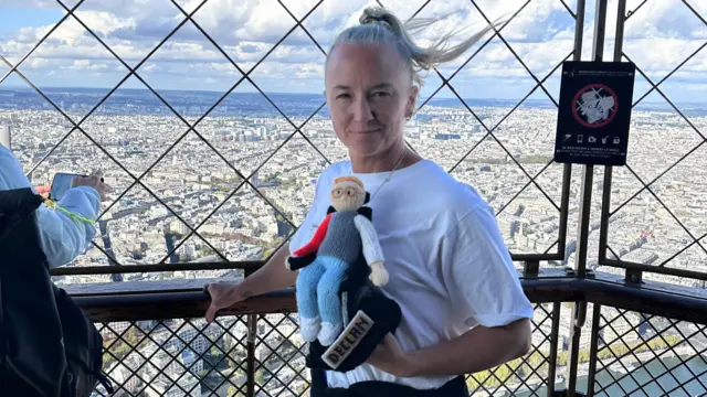 Alex está em pé na plataforma de observação da Torre Eiffel, segurando uma versão em tricô de Declan