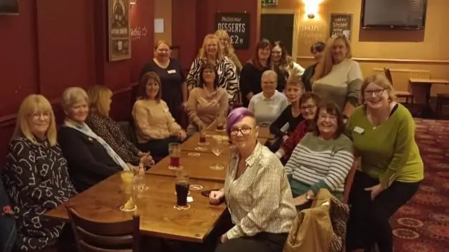 Um grupo de mulheres senta-se em torno de uma longa mesa em um pub. Elas estão olhando para a câmera e sorrindo. 