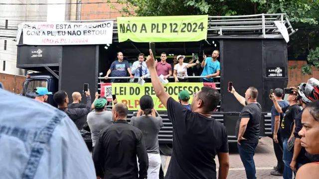 Manifestantes em frente a carro de som em protesto de motoristas contra projeto de lei, com faixas onde se lê "Se a Uber está de acordo com a PL, eu não estou!", "Não à PL 12/2024" e "Lei do Retrocesso PLP 12/2024"