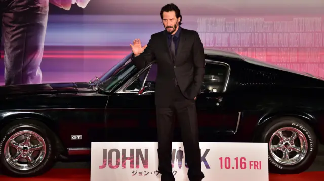 Keanu Reeves, vestindo calça e terno pretos, acena para a foto com um carro preto no fundo