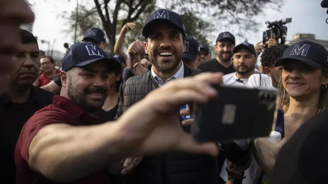 A foto mostra Marçal, um homem de barba cerrada, sorrindo, rodeado por pessoas, uma delas fazendo uma selfie com ele. Todos estão com um boné azul com a letra M. 