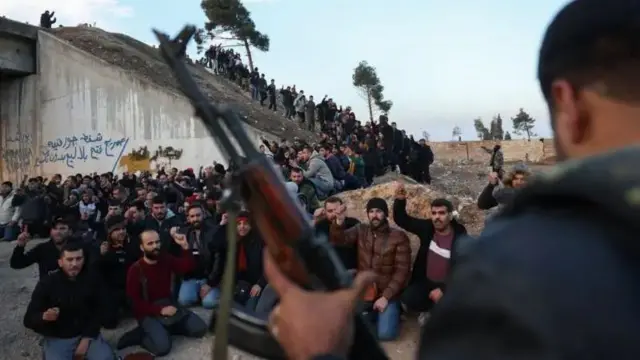 Foto de vários homens sentados ao redor de um outro homem que segura uma arma e está de pé.