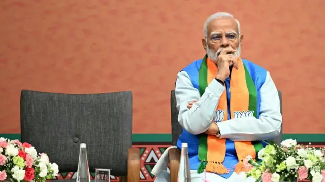 Narendra Modi looks thoughtful sitting at a table covered in flowers