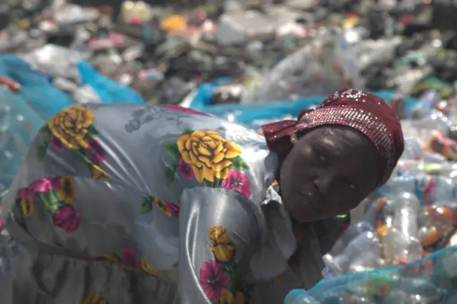 Abiba Alhassan, uma mulher de vestido florido e lenço na cabeça, separando garrafas plásticas usadas perto do lixão de Agbogbloshie, em Acra