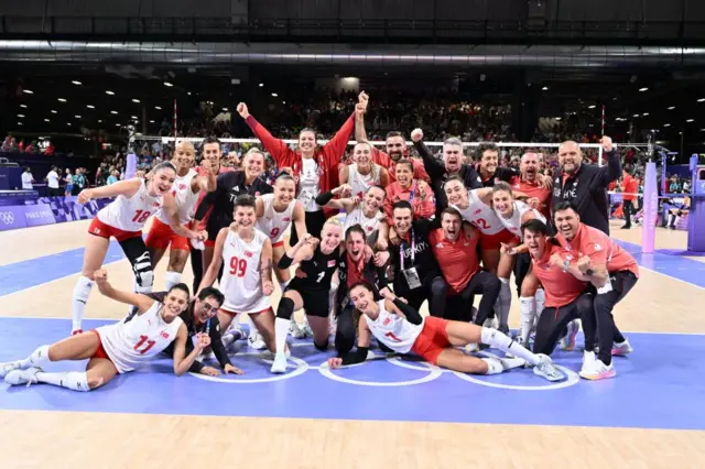 Kadın voleybol oyuncuları sahada hep birlikte poz veriyor. 