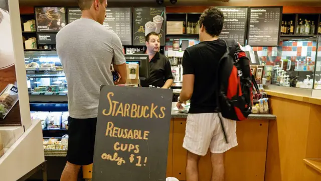 Dois clientes fazendo pedido no caixa de uma unidade da Starbucks, onde se vê uma placa oferecendo a venda de copos reutilizáveis por 1 dólar