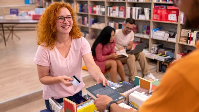 Jovem entregando livro e segurando cartão de crédito dentro de livraria