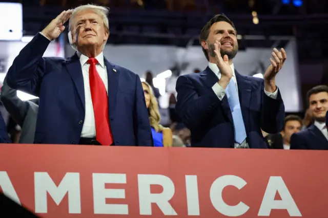 Trump e JD Vance em pé, sorrindo, durante grande evento, rodeados por outras pessoas