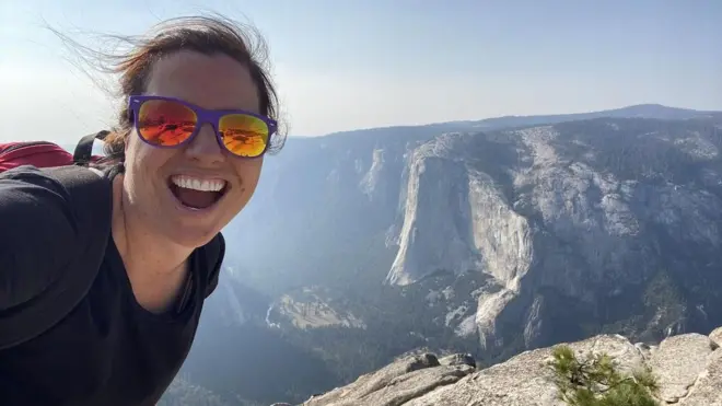 A advogada americana Jamie, que usa o pseudônimo M. E. Thomas, uma mulher branca jovem de óculos escuros espelhados, numa selfie sorrindo em uma paisagem montanhosa