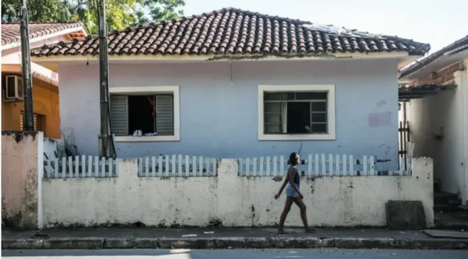 Mulher caminha em calçada à frente de casa azul, com duas janelas e muro