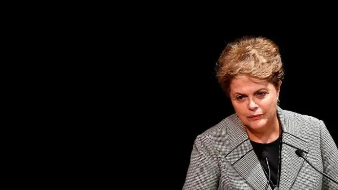 Dilma com olhar um pouco sério em pé, diante de microfone e fundo preto