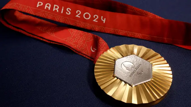 A foto mostra uma medalha dourada, com o centro em prateado escrito Paris 2024. Seu cordão é vermelho e também vem com o escrito Paris 2024.