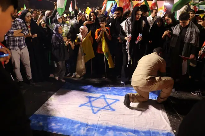 Em foto noturna, homem aparece ajoelhado em bandeira de Israel, observado por várias pessoas