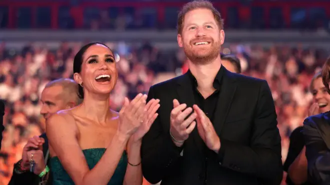 Jovem casal, uma mulher de pele morena e um homem branco; ambos sorriem e aplaudem