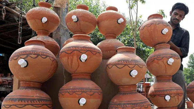 Potes de barros, com uma torneira no meio, empilhados. No canto à direita, um homem moreno, de cabelos curtos e camisa escura coloca um pote sobre o outro