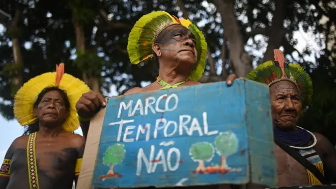 Três homens indígenas de meia idade durante protesto; um segura uma placa escrito 'marco temporal não'