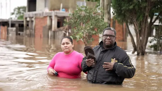 Rosana e Nelson com água até a cintura e roupas de frio em uma rua alagada